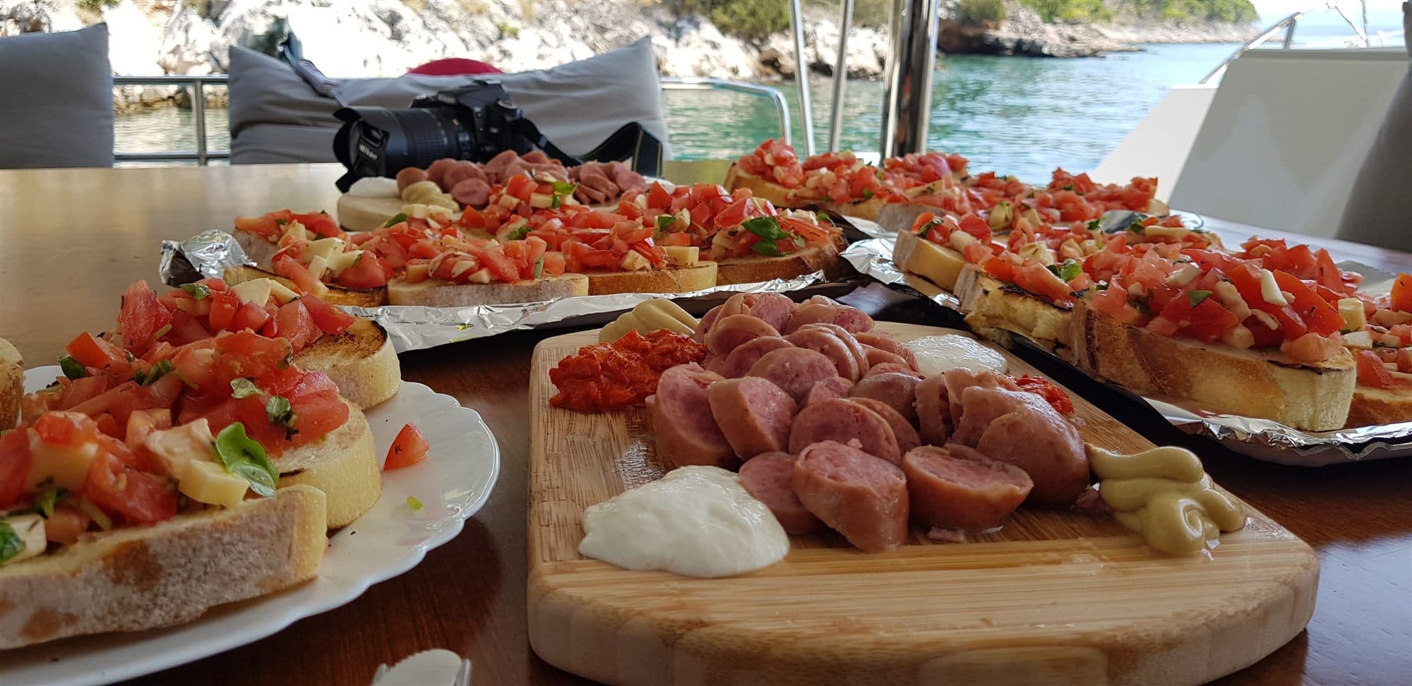 Lunch laid out on a yacht's cockpit table in a Croatian bay: boards of bruschetta with tomato and cheese, sliced sausage and dips, with the rocky shore and clear water behind