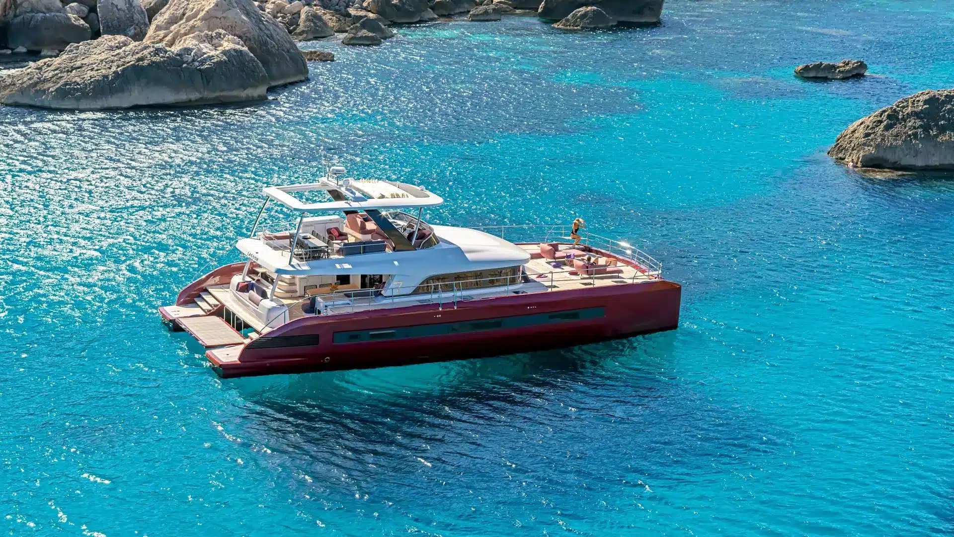 A large red-hulled power catamaran at anchor off pale rocks