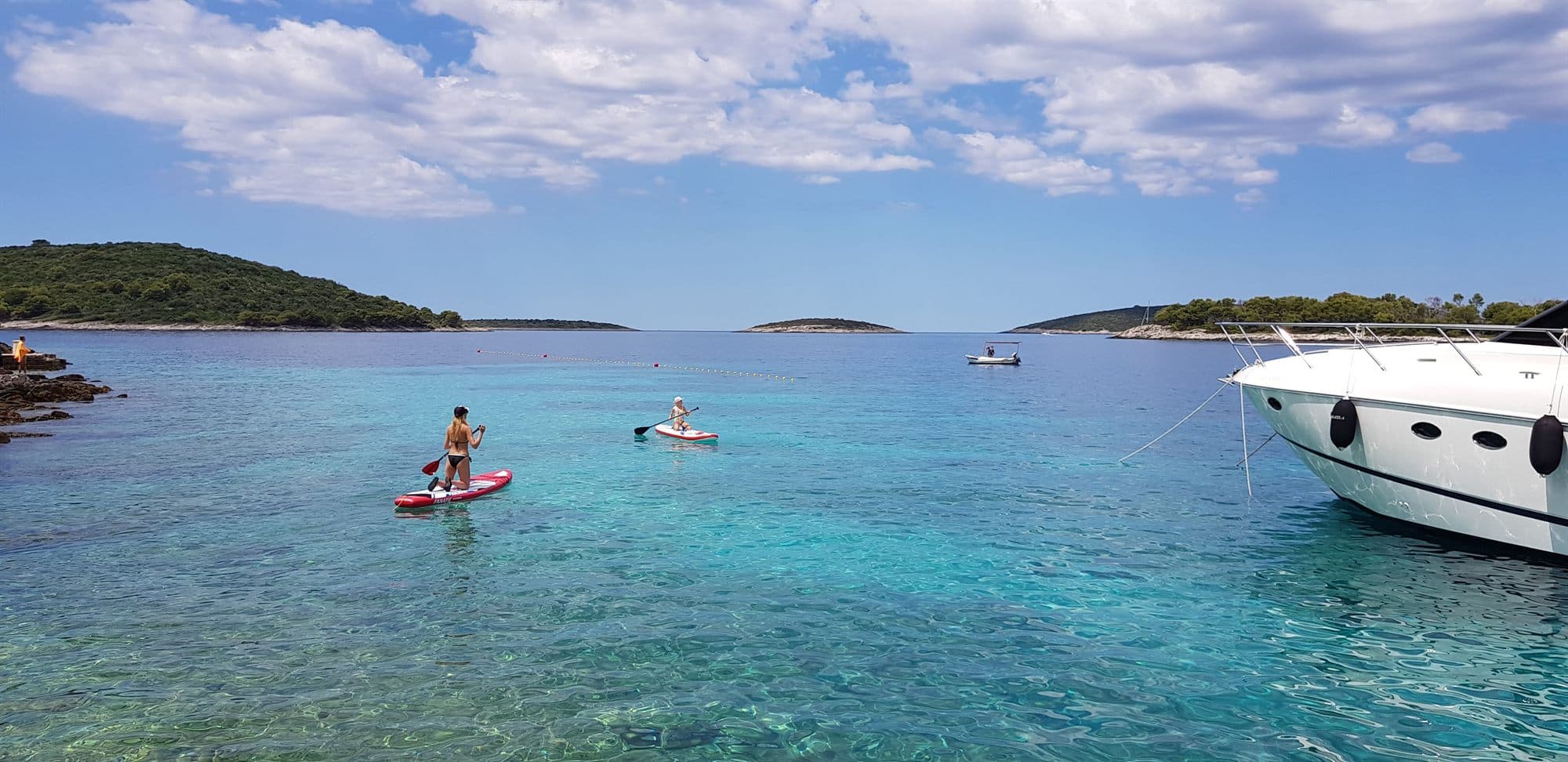 Two paddleboarders crossing a shallow turquoise Croatian bay, low islets on the horizon and a motor boat moored at the right