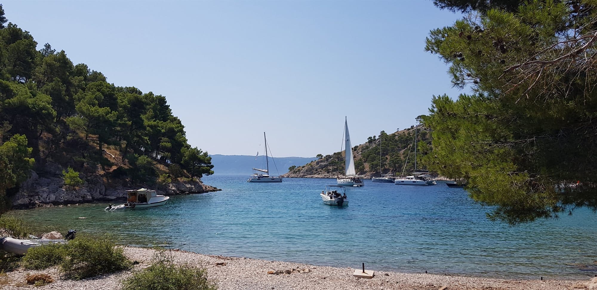 A sheltered Croatian cove framed by pines, a yacht anchored in clear turquoise water and a pebble beach in the foreground