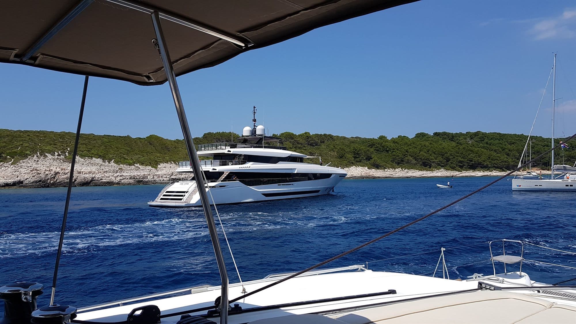 A large white superyacht anchored close in against a pine-covered Croatian island, sailing yachts moored beyond her in the same bay